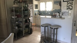 A kitchen with a metal shelving unit, two stools, a refrigerator, and a window with curtains. - VILLA WITH POOL, PANORAMIC VIEW, 5 MINUTES WALK FROM THE VILLAGE (Lorgues)