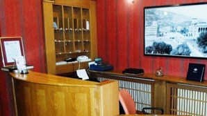 A reception desk with a wooden counter, a glass display case, a framed photograph on the wall, and a round table with a placemat. - Hotel Skopje (Skopje)