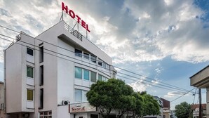 A hotel with a sign reading 'HOTEL' on the top, a storefront with 'SKOPJE' signage, and parked cars along the street. - Hotel Skopje (Skopje)