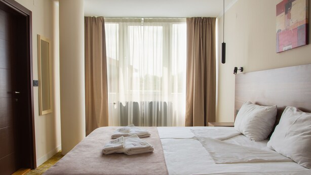 A hotel room with a neatly made bed, a door, a window with curtains, and a painting on the wall. - Hotel Skopje (Skopje)