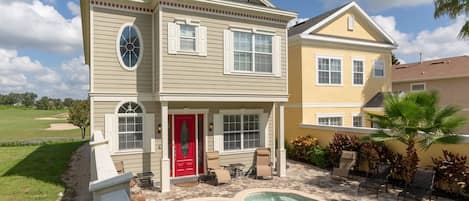 A two-story house with a swimming pool, a red door, and a patio with chairs and a table.
