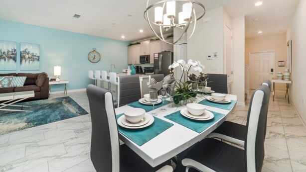 A modern dining area with a round table set for four, a kitchen in the background, and a living room with a sofa and artwork. - Rent a Luxury Townhome on Storey Lake Resort, Minutes From Disney, Orlando Townhome 1004 (Kissimmee)