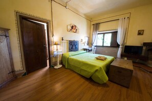 Una stanza con pavimento in legno, un letto con copriletto verde, un armadietto in legno, una lampada, un quadro appeso al muro e una finestra con tende.