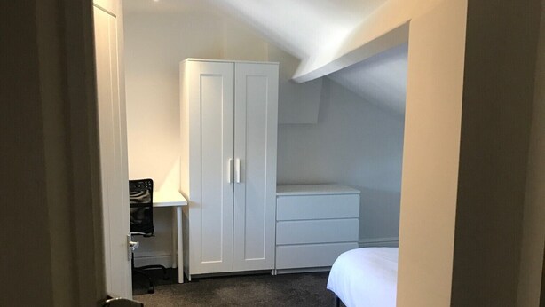 Een kleine, zwak verlichte kamer met een enkel bed, een witte kast en een bureau met een stoel.