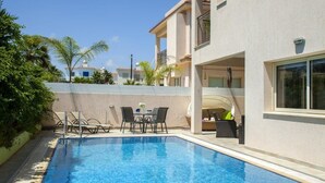 A swimming pool with lounge chairs and a table, surrounded by a building with balconies and a clear sky. - Beautiful Villa With Private Pool, Protaras Villa 1028 (Paralimni)