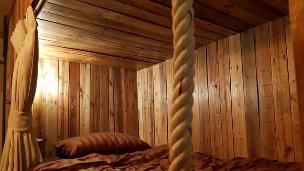 A wooden cabin-style room with a bed, a rope, and a wooden ceiling. - Cozy Cabin - Hostel (Bangkok)