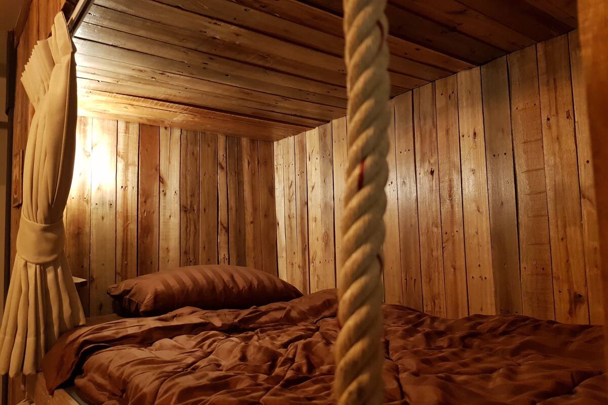 A wooden cabin-style room with a bed, a rope, and a wooden ceiling.