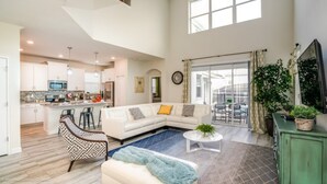 A spacious living area with a white sofa, a patterned armchair, and a coffee table. A kitchen with white cabinets and a dining area with a table and chairs are visible. - Luxury 5 Bedroom Villa on Champions Gate Resort, Orlando Villa 3112 (Davenport)