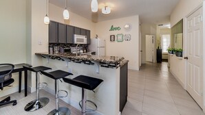 A modern kitchen with dark cabinets, a granite countertop, and a central island with bar stools. There are pendant lights above the island and a microwave on the countertop. - Luxury 3 Bedroom House on Windsor Hills Resort, Orlando House 1900 (Kissimmee)