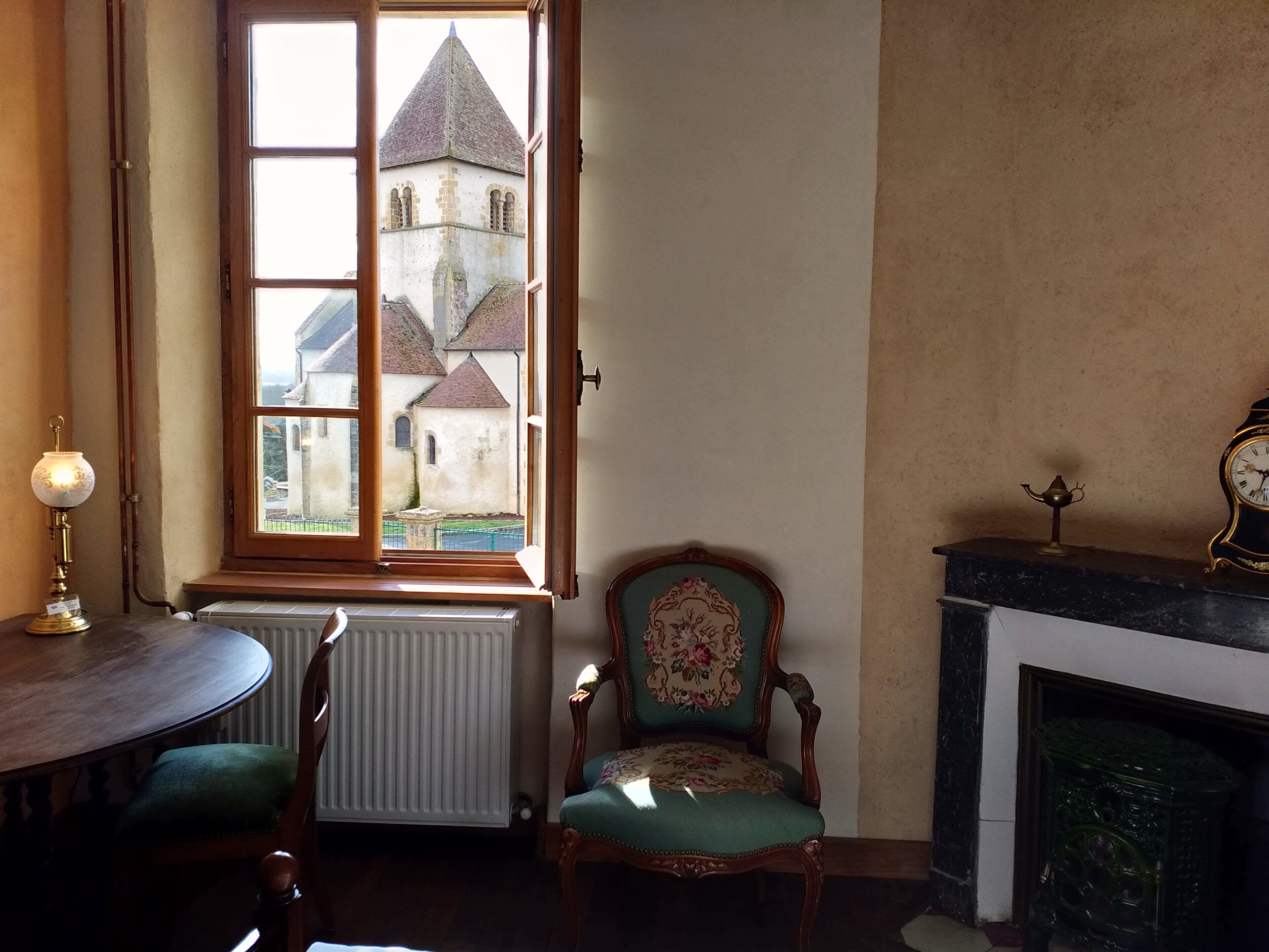 Habitació amb vista a una església a través d'una finestra, un radiador, una cadira tapissada de verd, una taula i una llar de foc amb un rellotge.