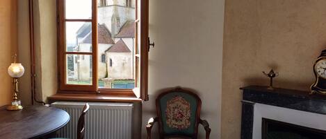 Een kamer met uitzicht op een kerk door een raam, een radiator, een groene bekleden stoel, een tafel en een open haard met een klok.