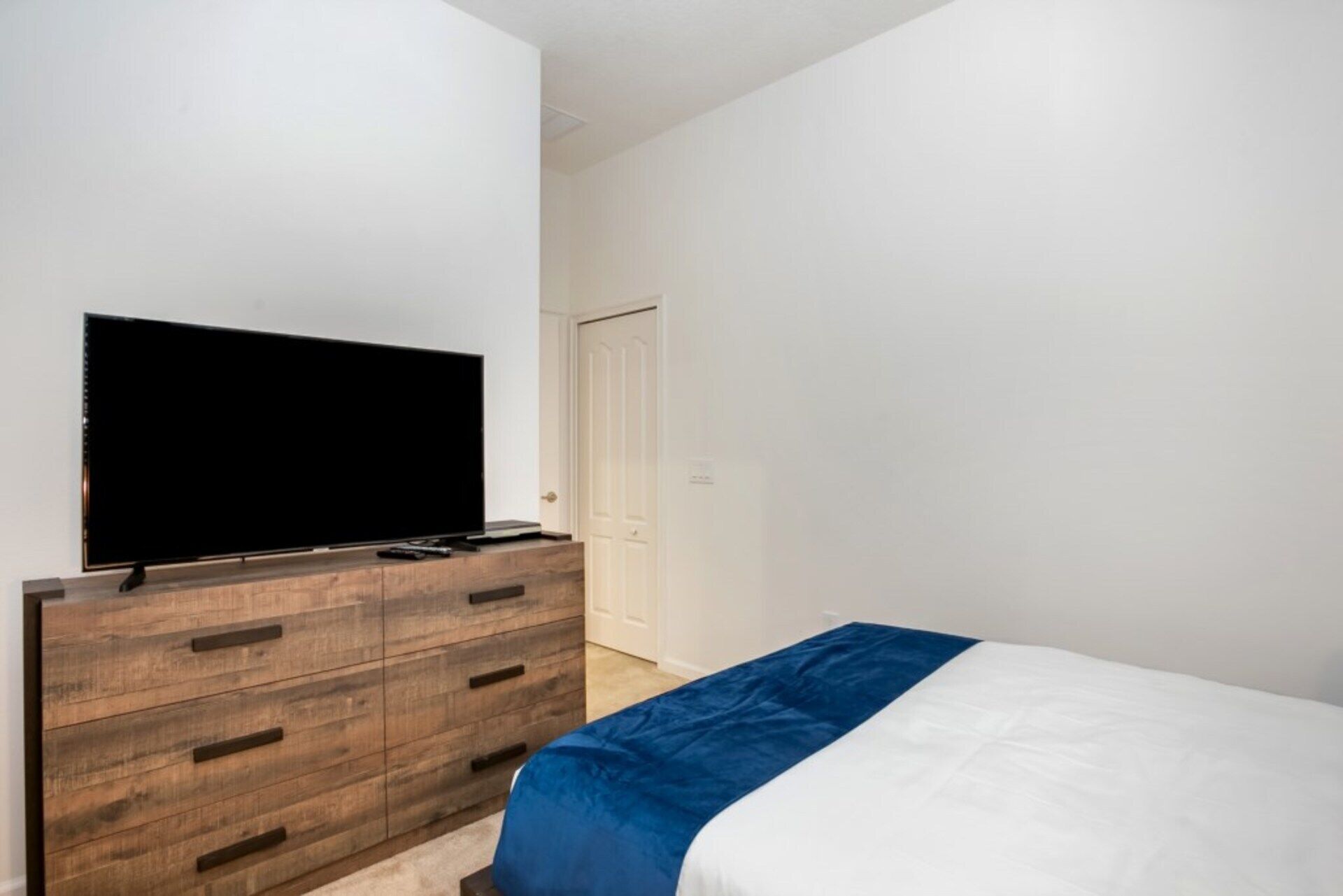 A bedroom with a wooden dresser, a flat-screen TV, a bed with a blue and white bedspread, and a white door.