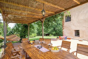 A wooden table with chairs, a hanging hammock, and a table set with wine glasses and fruit.