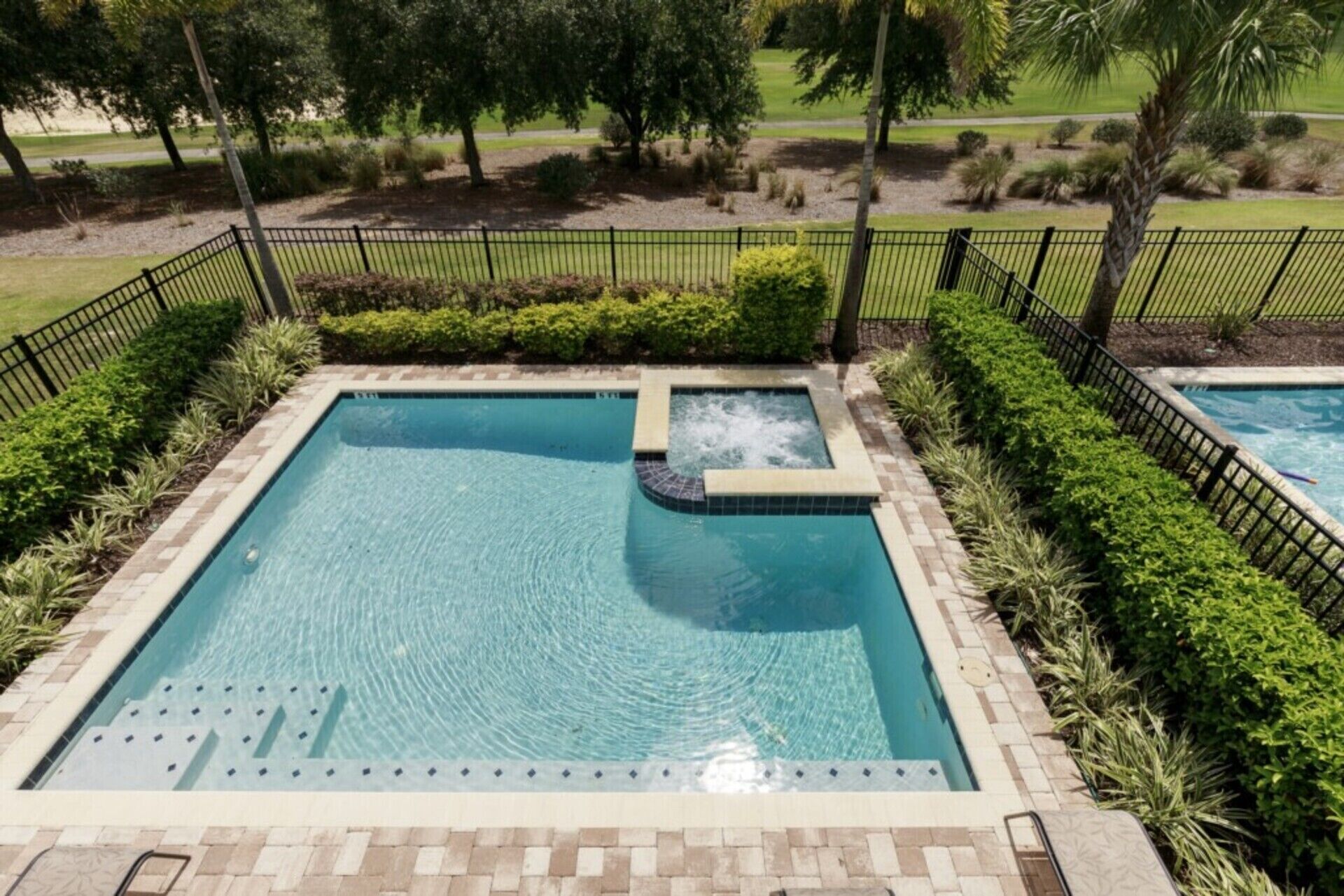 A rectangular pool with a hot tub, surrounded by a black metal fence and lush greenery.