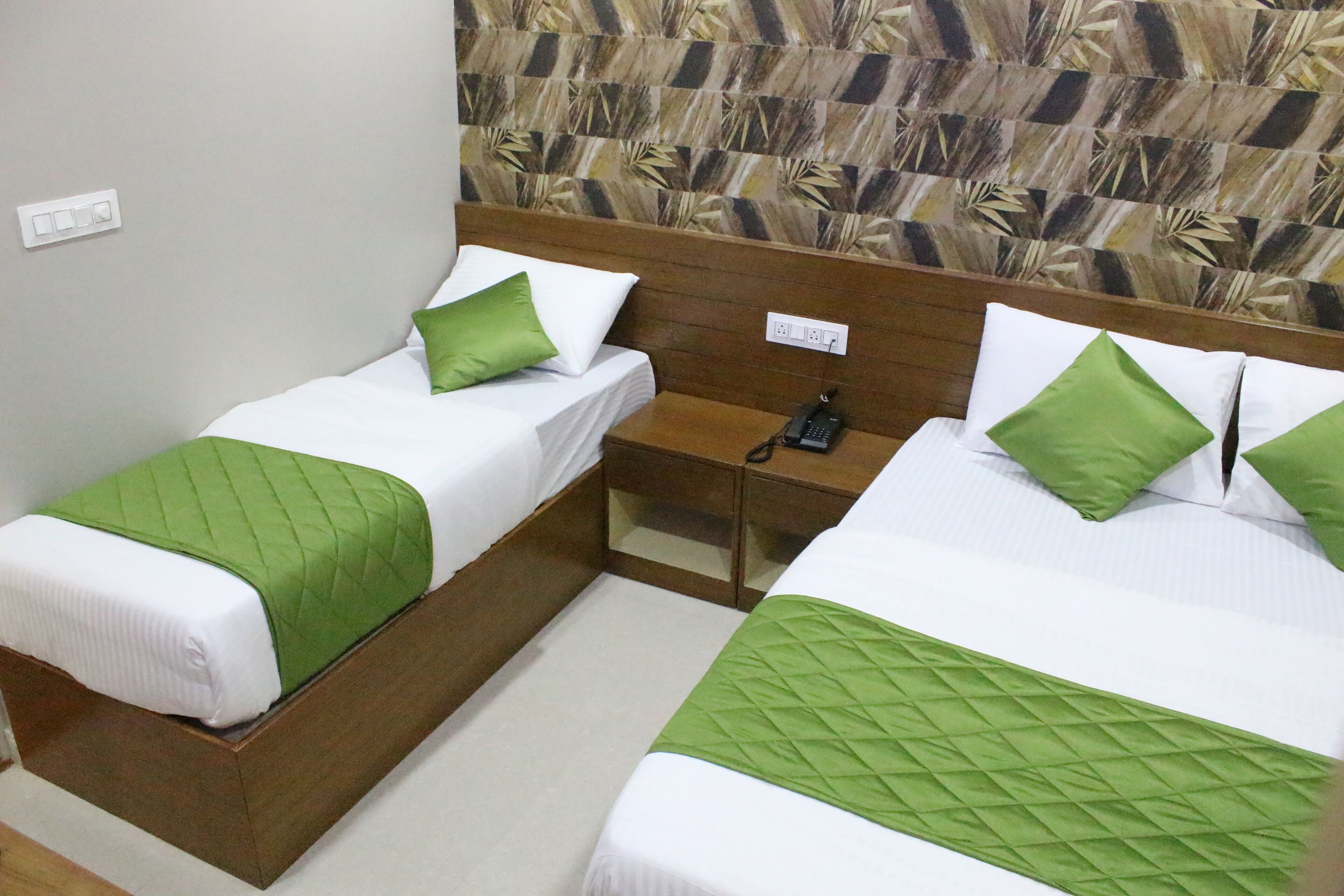 A hotel room with two single beds, each with a green quilted bedspread and matching pillows. There is a wooden nightstand between the beds with a black telephone. The room has a patterned wallpaper on the wall behind the beds.