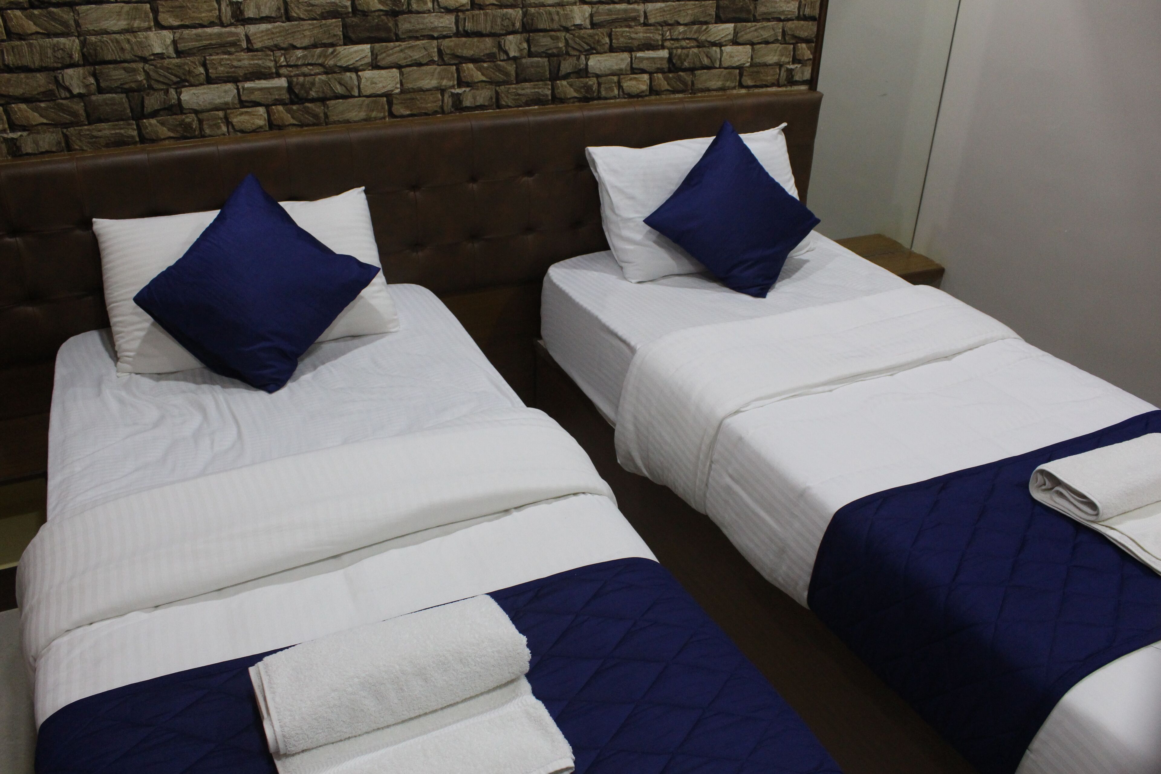 Two twin beds with white linens and blue pillows, a brown headboard, and a stone wall in the background.
