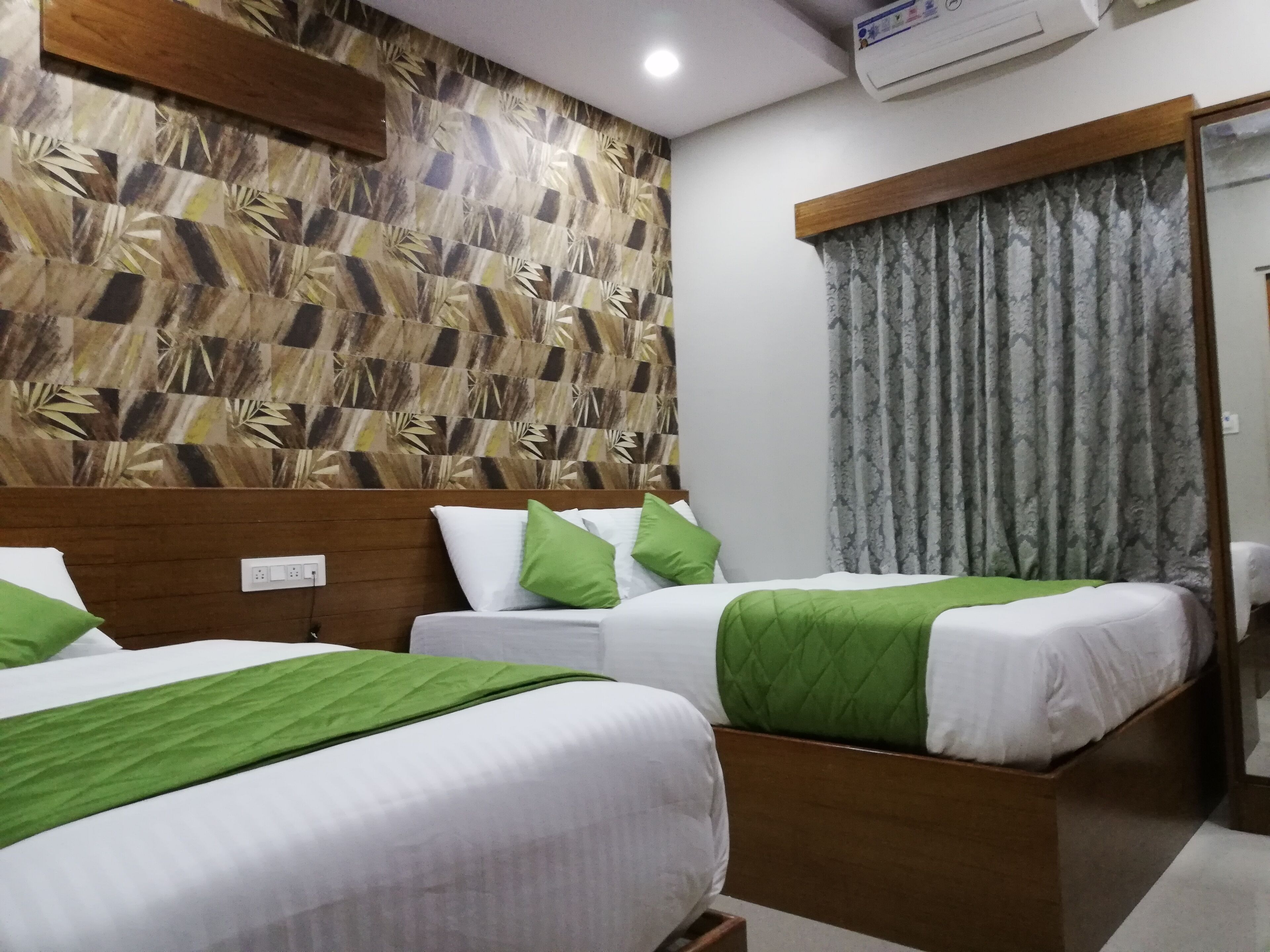 A hotel room with two single beds, each with a green bedspread and pillow, a wooden headboard, and a patterned wall.