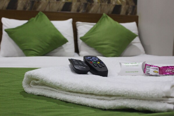 A neatly made bed with green pillows, two remote controls, and a bar of moisturizing soap on a green towel.