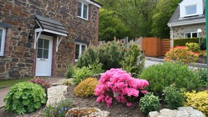 Uma casa de pedra com porta branca, rodeada por um jardim com diversas plantas e flores.