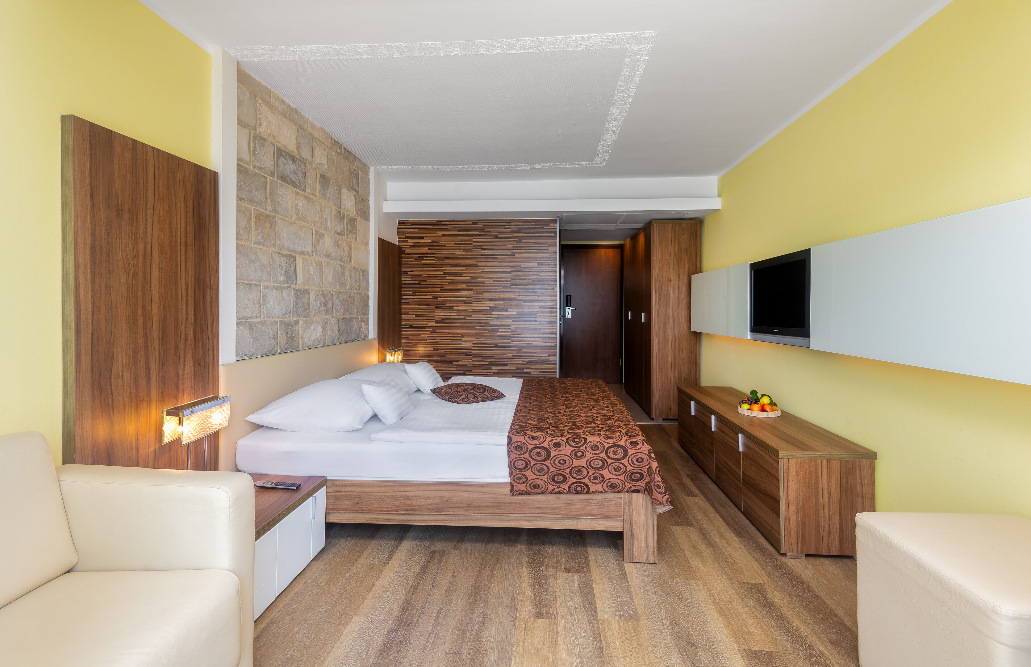 A modern hotel room with a wooden bed, a flat-screen TV mounted on the wall, and a wooden nightstand with a lamp.