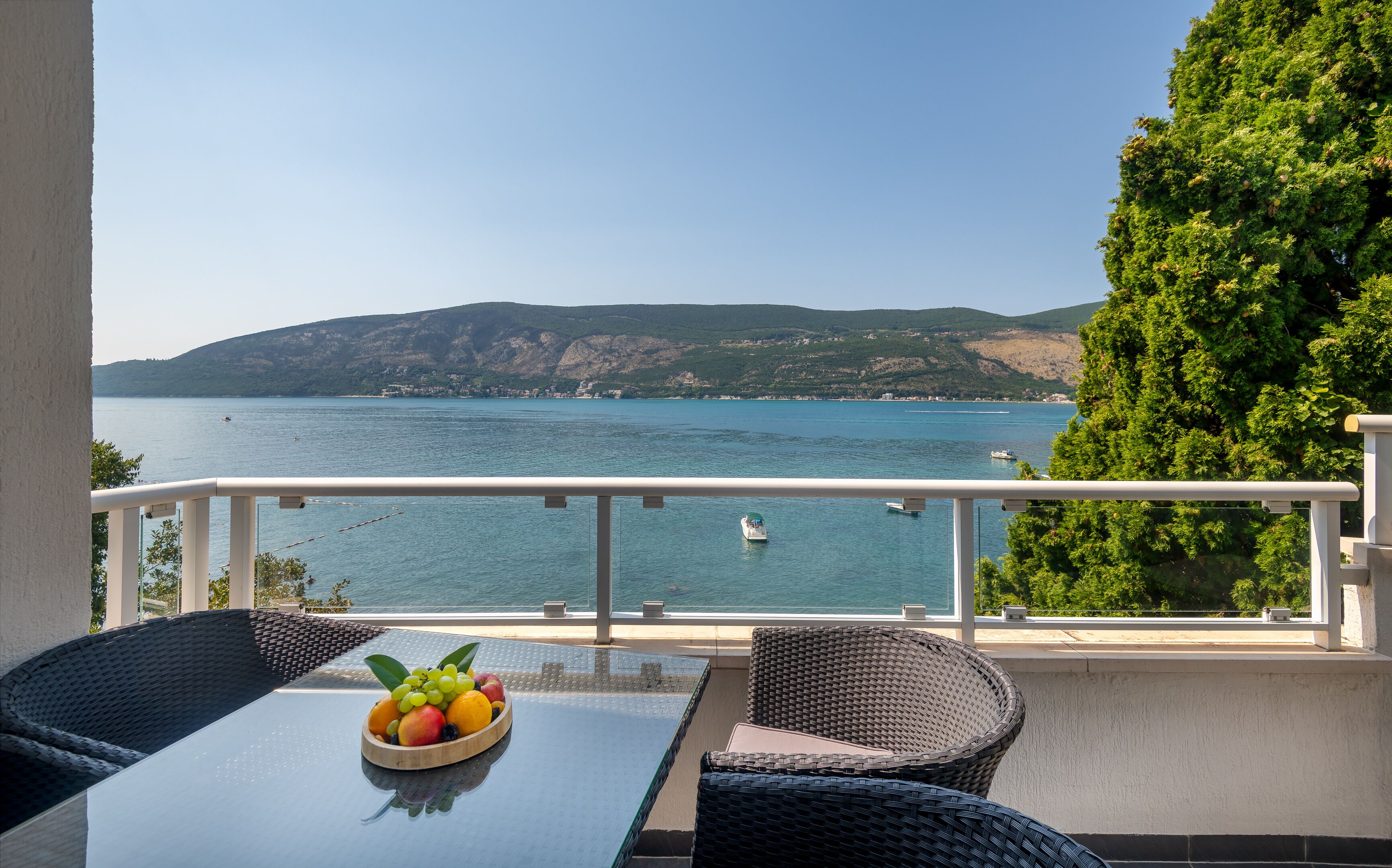 Un balcón con una mesa y sillas, un frutero y vistas a un lago con barcos y colinas.
