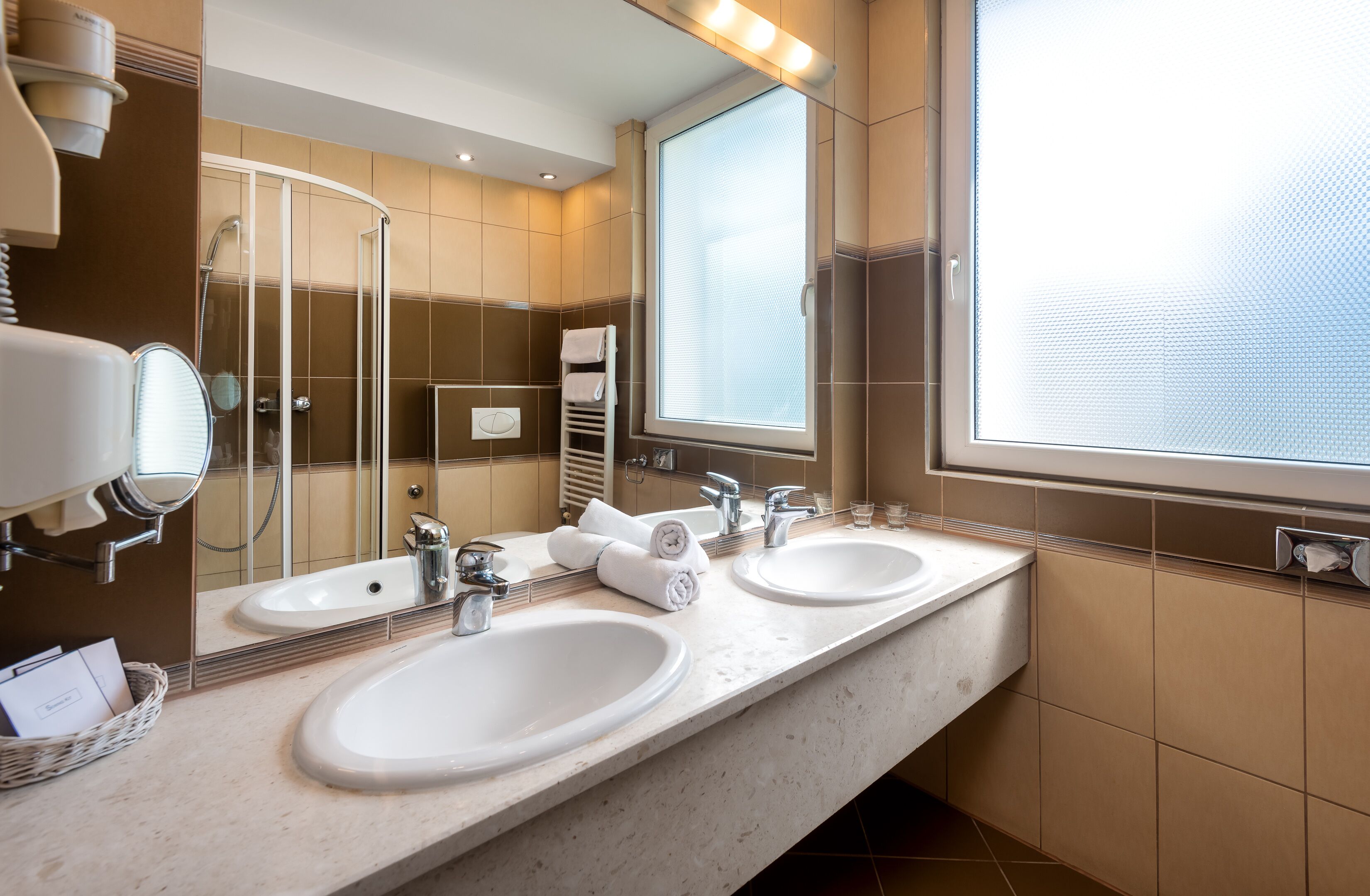 A hotel bathroom with two sinks, a large mirror, a shower, and a window.