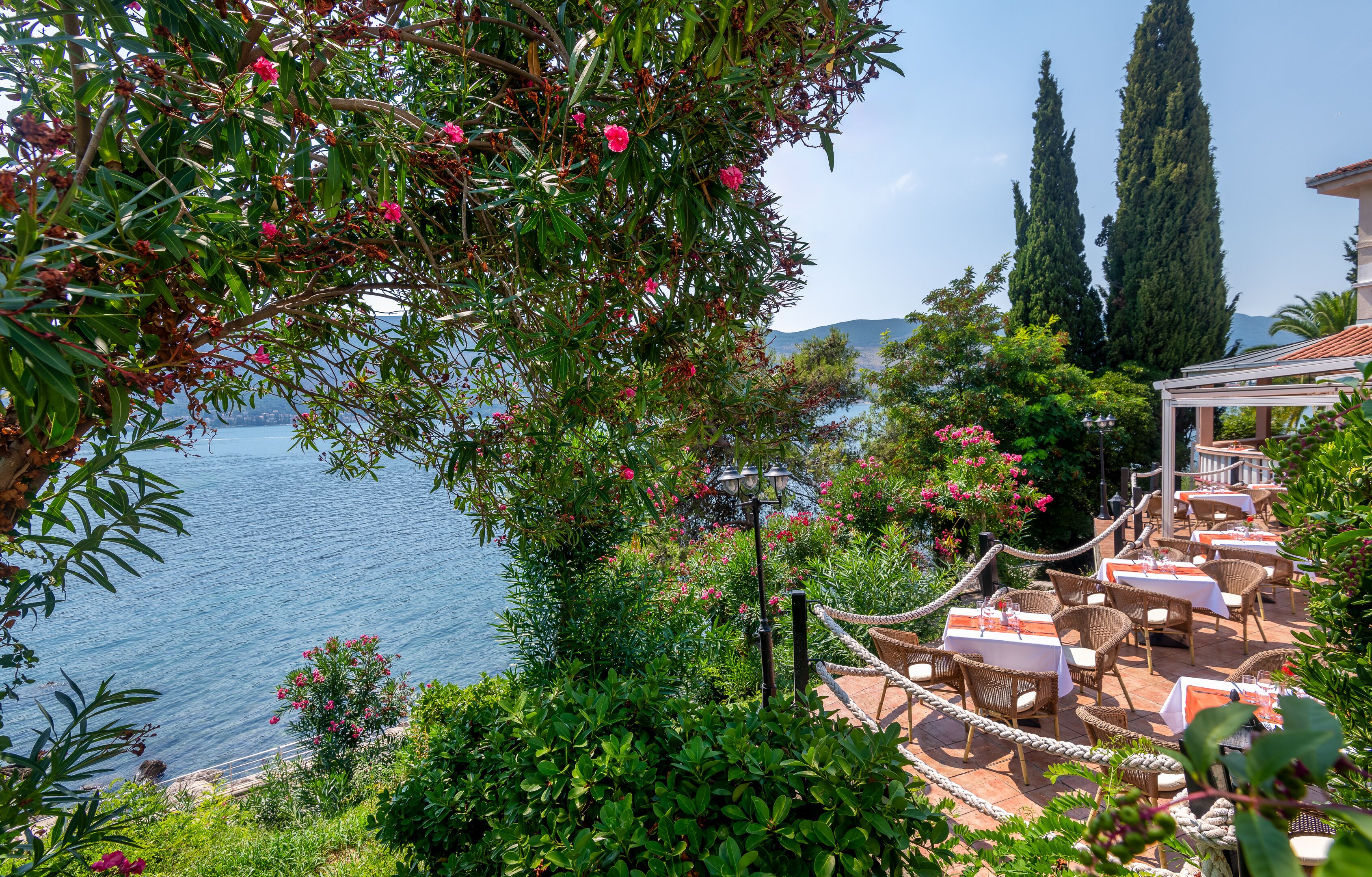 Un espace de restauration en plein air situé en bord de mer, avec des meubles et des tables en osier, offrant une vue sur l’eau et la végétation environnante.