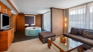 Ein modernes Hotelzimmer mit einem Flachbildfernseher, einem großzügigen Sitzbereich mit einem Glastisch und einer runden Whirlwanne sowie einem großen Fenster mit Vorhängen.