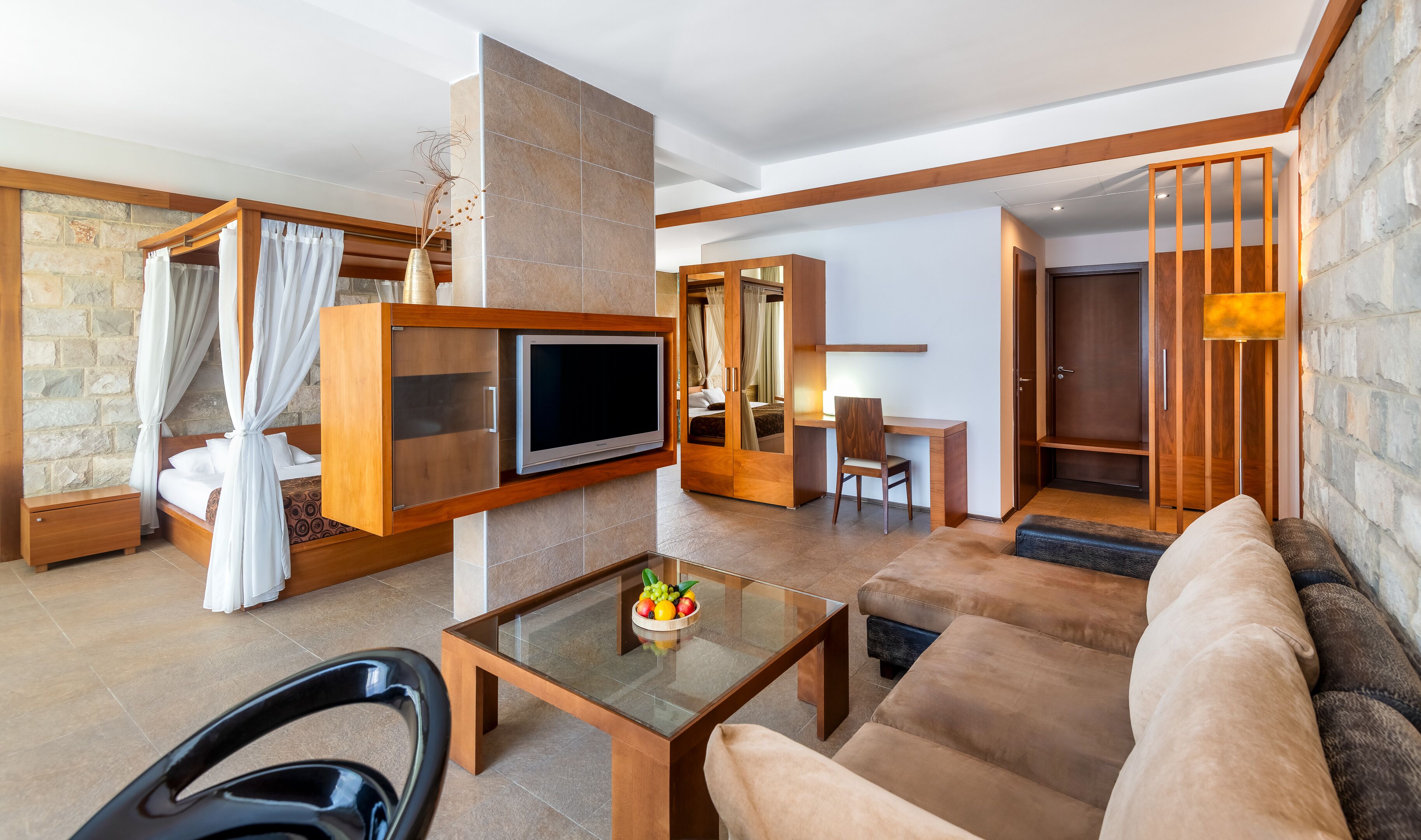 Una habitación de hotel moderna con televisión, una mesa de centro de vidrio con fruta, un sofá, un escritorio y una cama con cortinas.