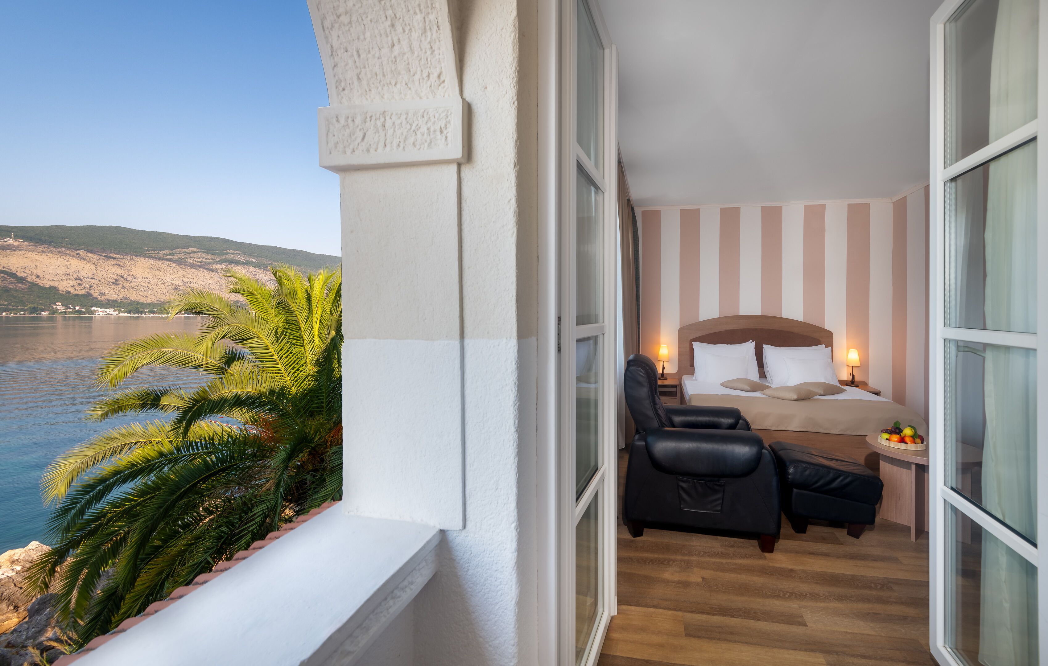 A balcony with a view of the sea, a bed with a headboard, a chair, and a small table with fruit.