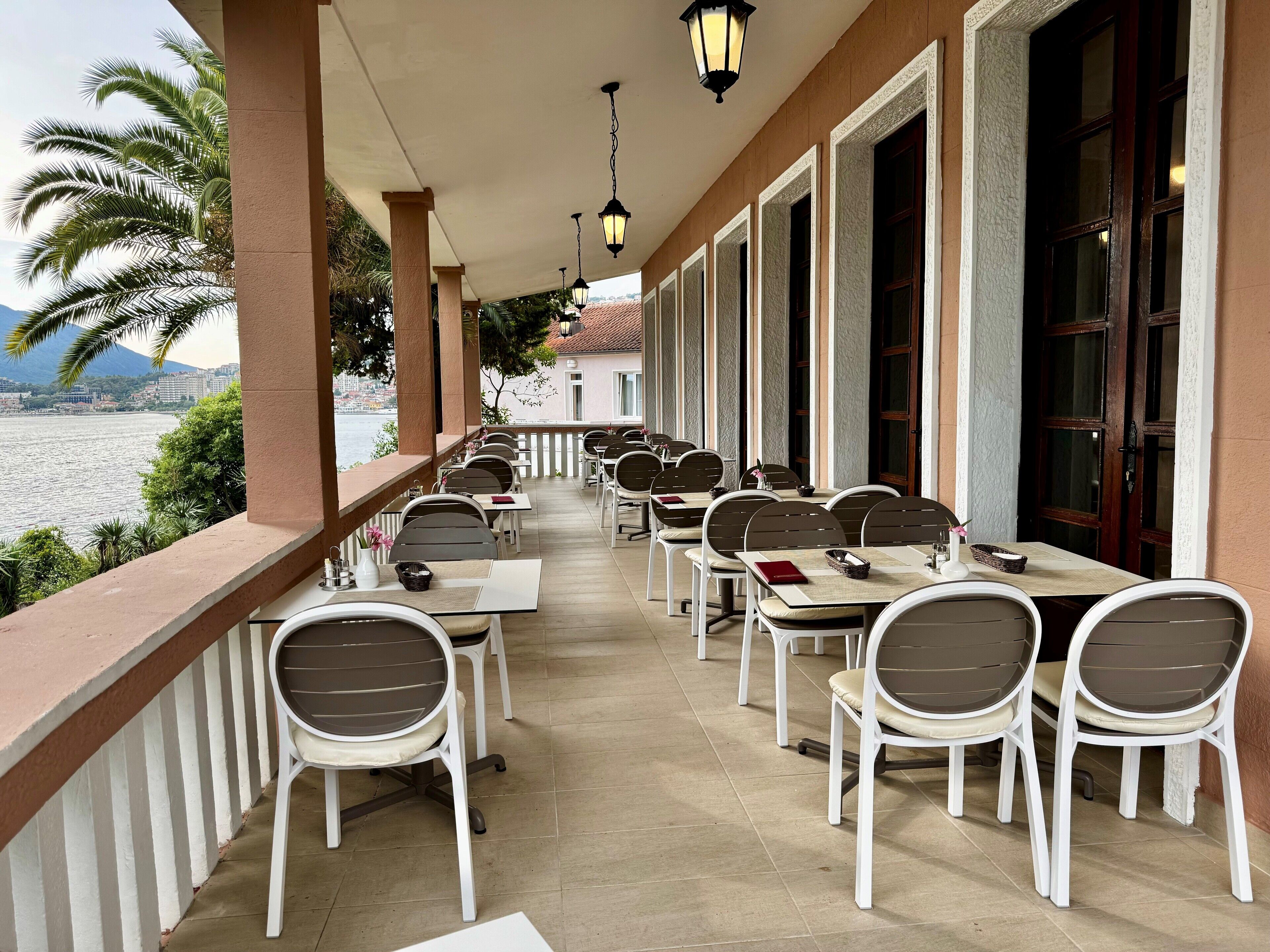 A covered outdoor dining area with tables and chairs, overlooking a body of water and mountains.