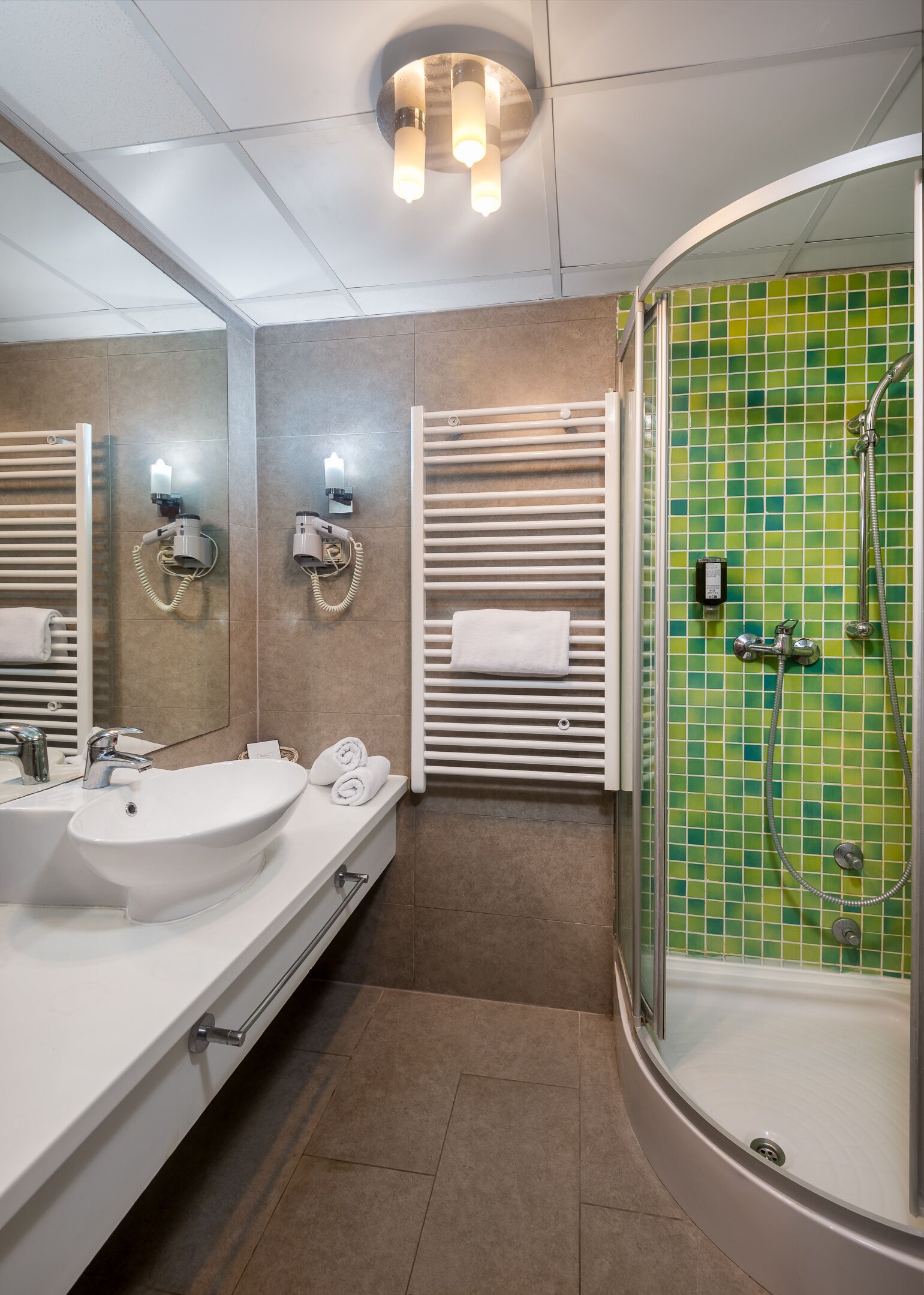 A modern bathroom with a large sink, two hair dryers, a towel rack, and a shower with a green mosaic wall.