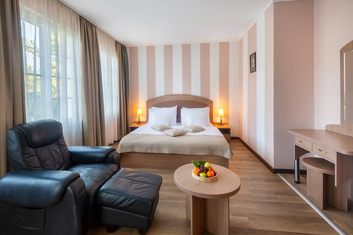 Ein Hotelzimmer mit einem Bett, einem Ledersessel, einem kleinen runden Tisch mit Früchten und einem Schreibtisch mit Schubladen.