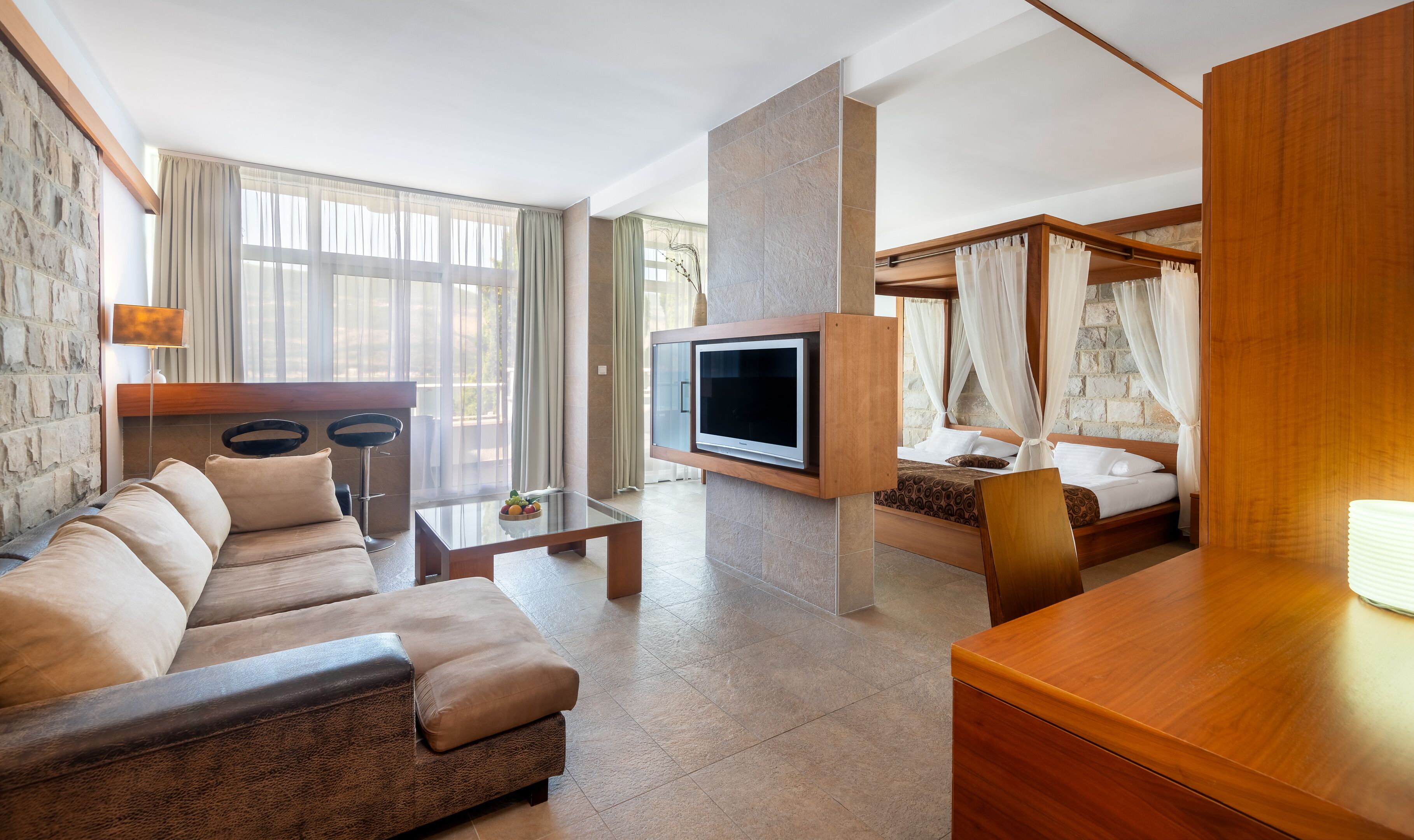 A modern hotel room with a sofa, a TV mounted on a wall, a bed with a canopy, and a stone accent wall.