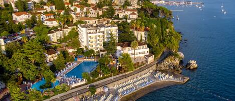 An aerial view of a coastal hotel with a swimming pool, beachside lounge chairs, and a view of the sea and mountains.