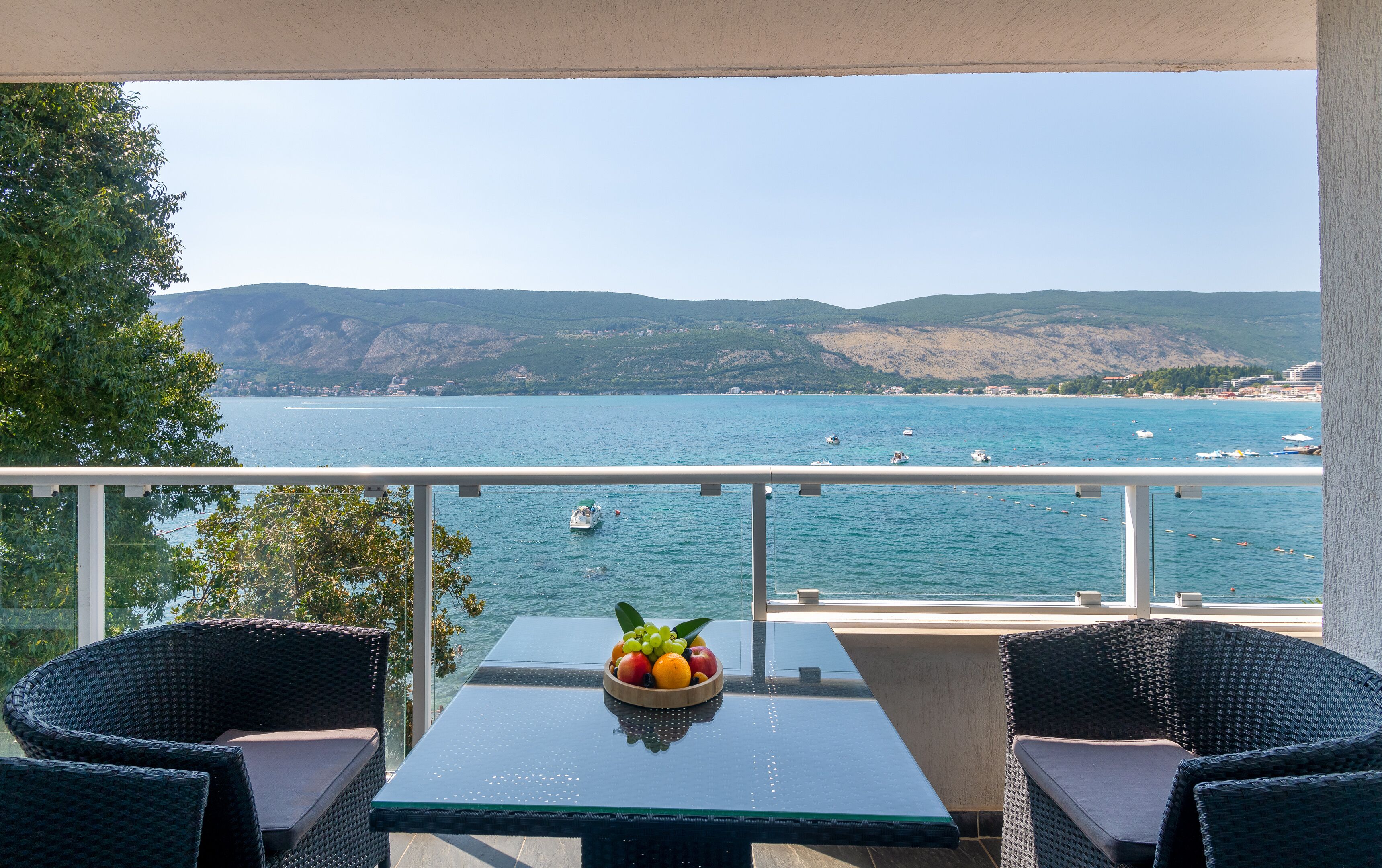 Un balcón con sillas de mimbre y una mesa, con vistas a la costa, barcos y colinas.