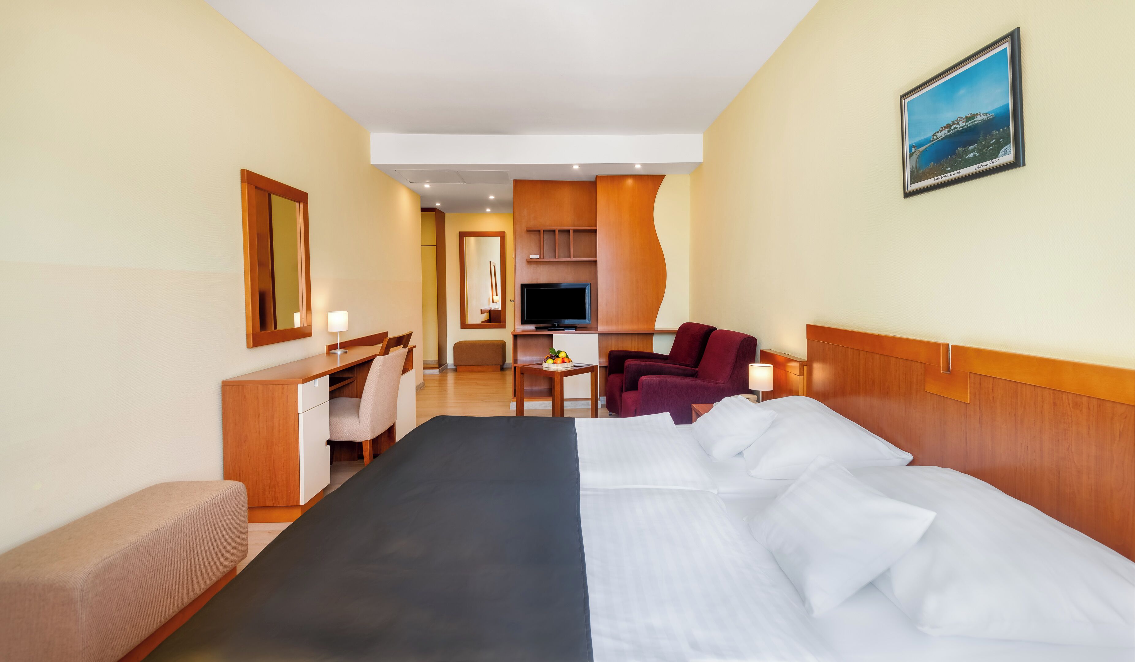 Une chambre d’hôtel avec un grand lit, un bureau avec une chaise, une télévision, une petite table avec des fruits et un fauteuil rouge.