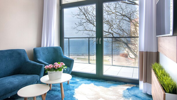 A room with a blue sofa, a small round table with a flower pot, and a view of the outdoors through a large glass door. - Apartament z widokiem na morze - Nadmorski Luksus Apartamenty (Ustronie Morskie)