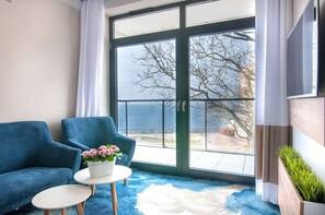 A room with a blue sofa, a small round table with a flower pot, and a view of the outdoors through a large glass door. - Apartament z widokiem na morze - Nadmorski Luksus Apartamenty (Ustronie Morskie)