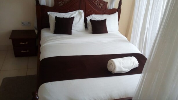 A neatly made bed with white and brown bedding, a wooden headboard, and a small bedside table with drawers. - MT Ararat Hotel (Nakuru)