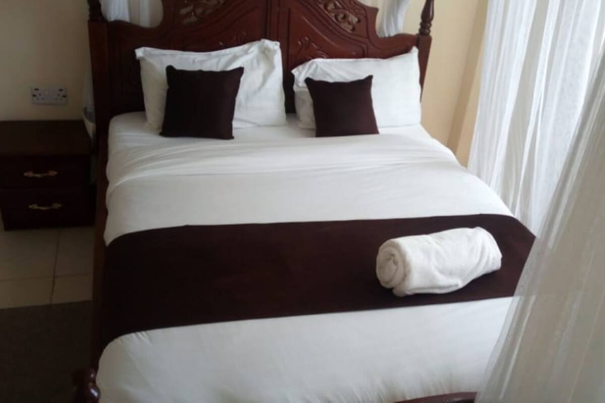 Una cama bien hecha con una colcha marrón y sábanas blancas, una mesita de noche de madera con un cajón y un enchufe en la pared.