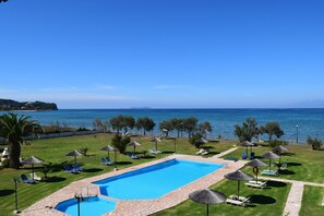 A swimming pool with lounge chairs and umbrellas, surrounded by a grassy area and a view of the sea. - S&C Kosmos Beach Resort (Corfu)