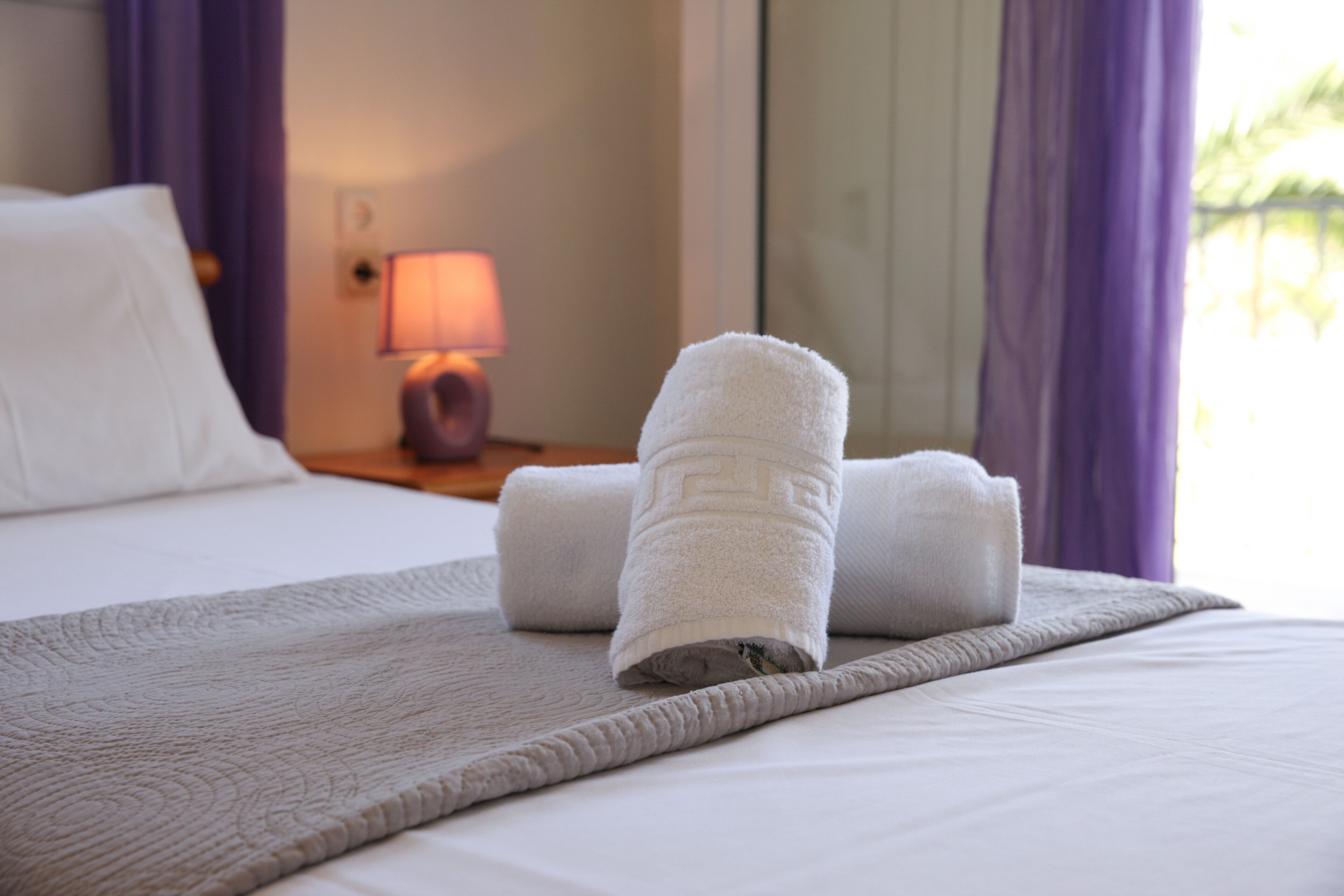A neatly made bed with rolled towels, a bedside lamp, and purple curtains.