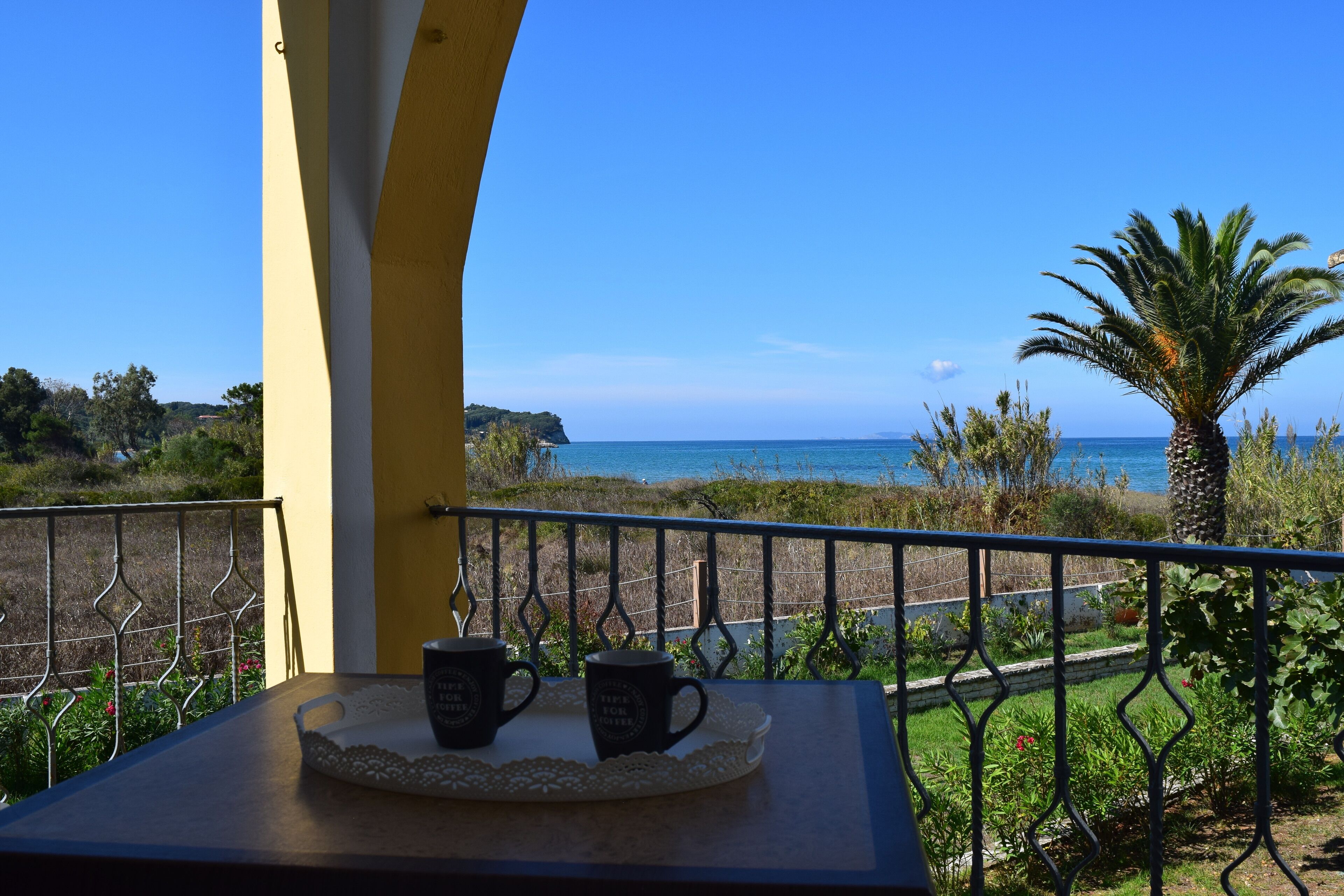 A balcony with two black mugs on a tray, overlooking a coastal landscape with a clear sky.