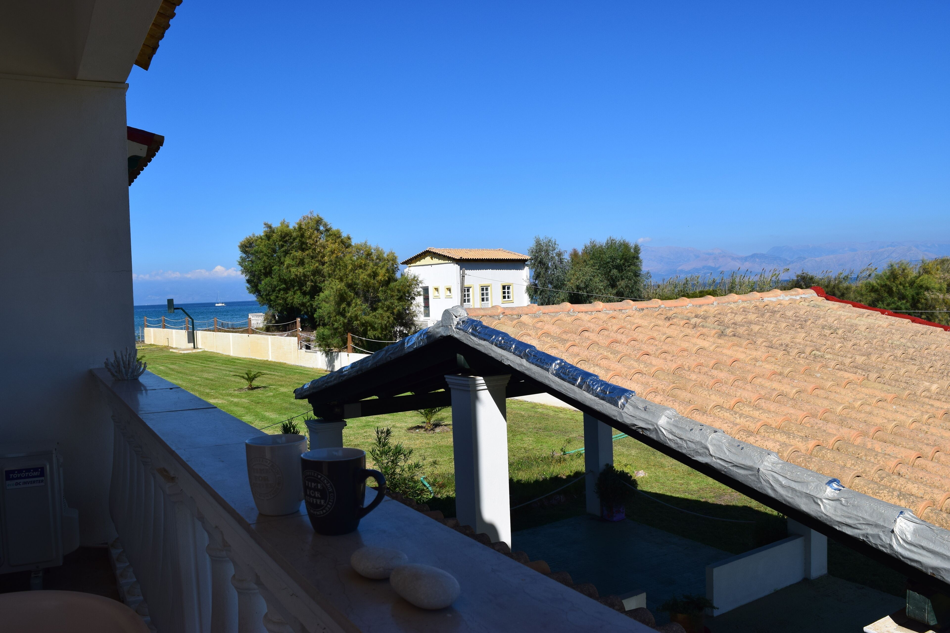 A balcony with a view of the sea, a tiled roof, and a white building in the background.
