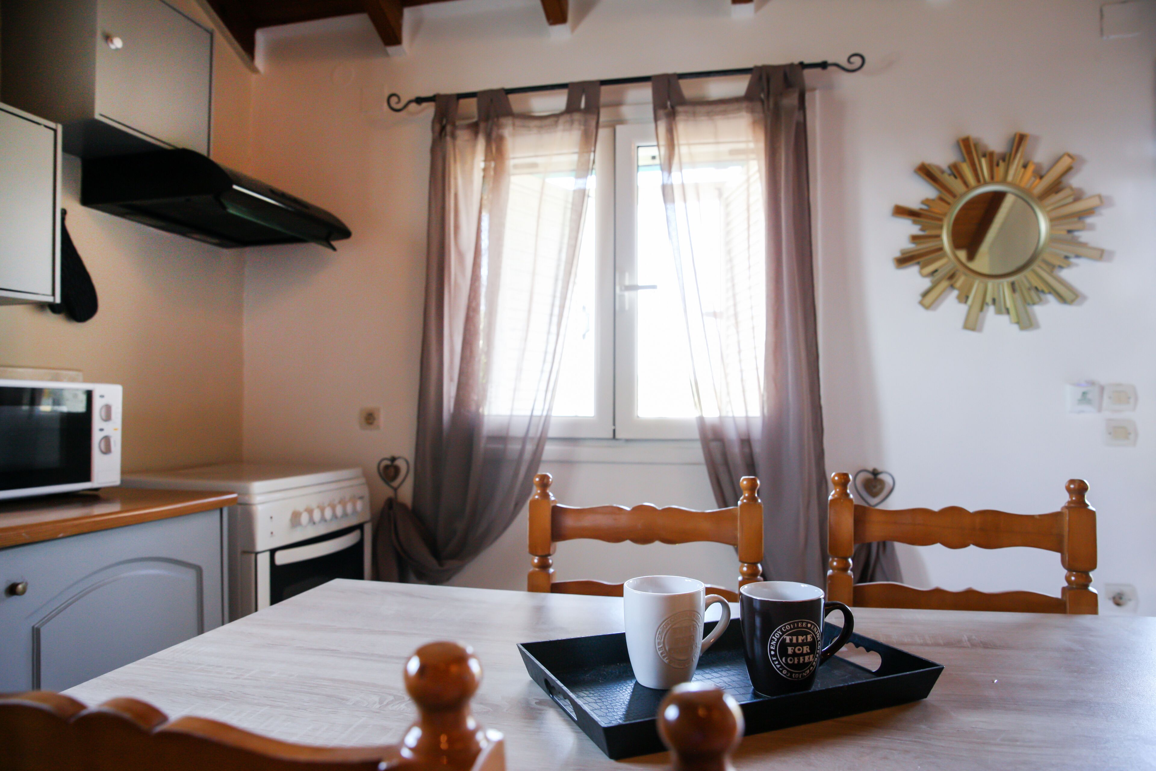 A kitchen with a microwave, stove, and wooden chairs. There is a window with curtains and a sunburst mirror on the wall.
