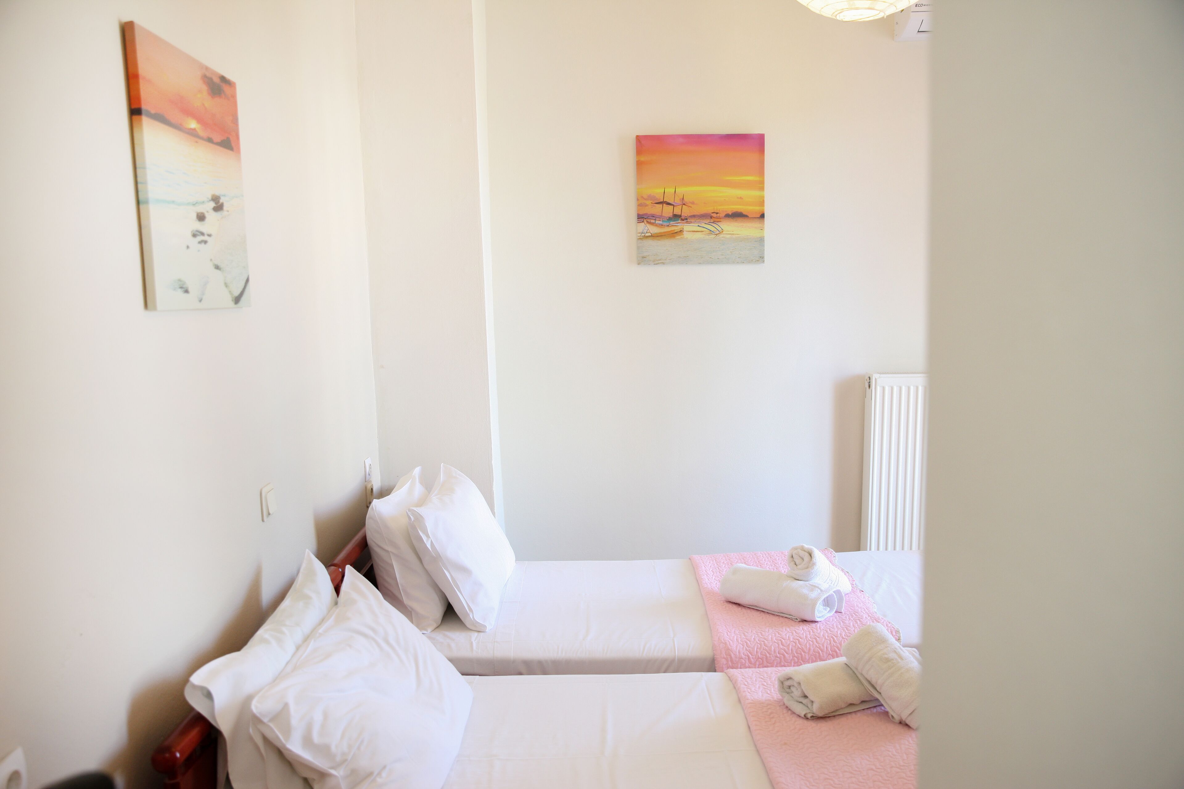 Een hotelkamer met twee bedden, witte beddengoed, een roze plaid en twee schilderijen aan de muur.