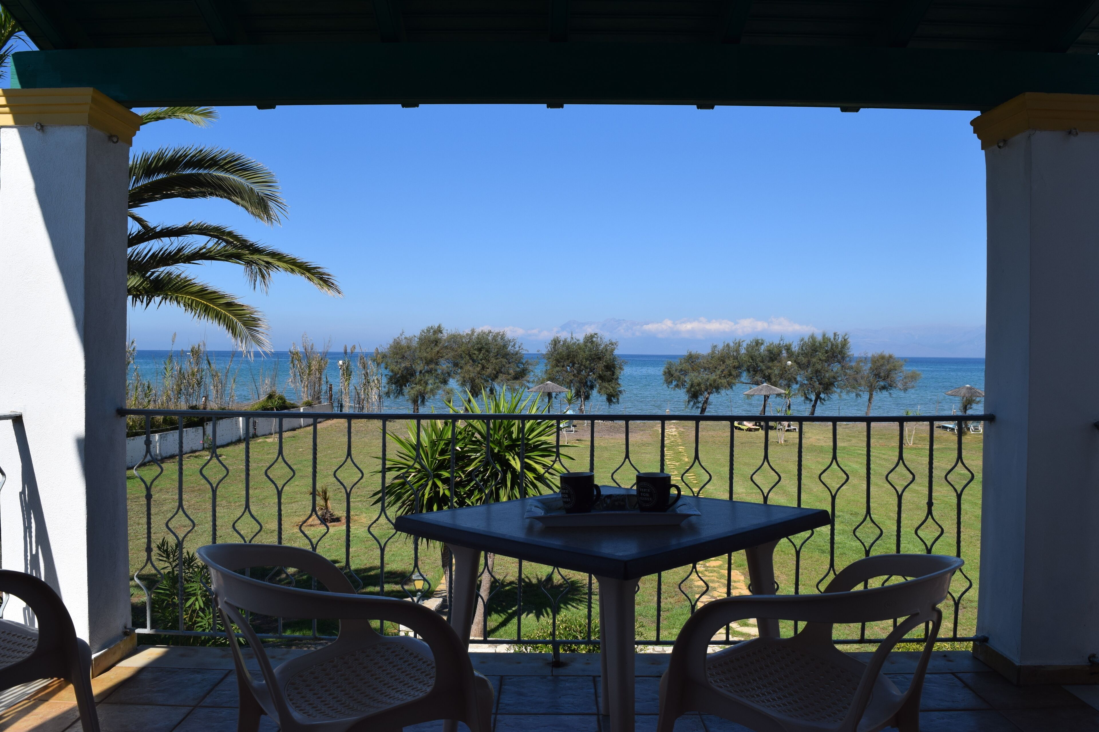A balcony with a table and chairs, overlooking a clear blue sea and a grassy area with trees.
