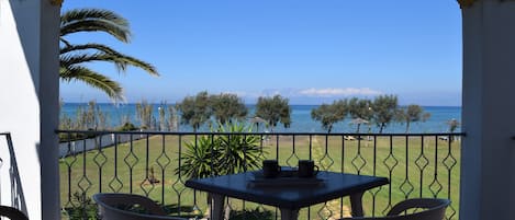 A balcony with a table and chairs, overlooking a clear blue sea and a grassy area with trees.