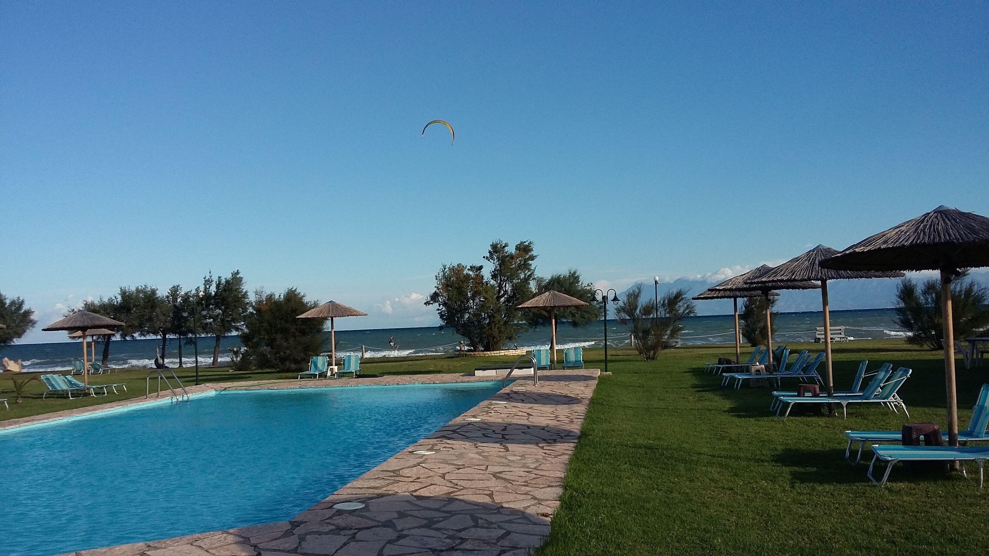 A pool area with lounge chairs and umbrellas, a clear view of the ocean, and a parasail in the sky.